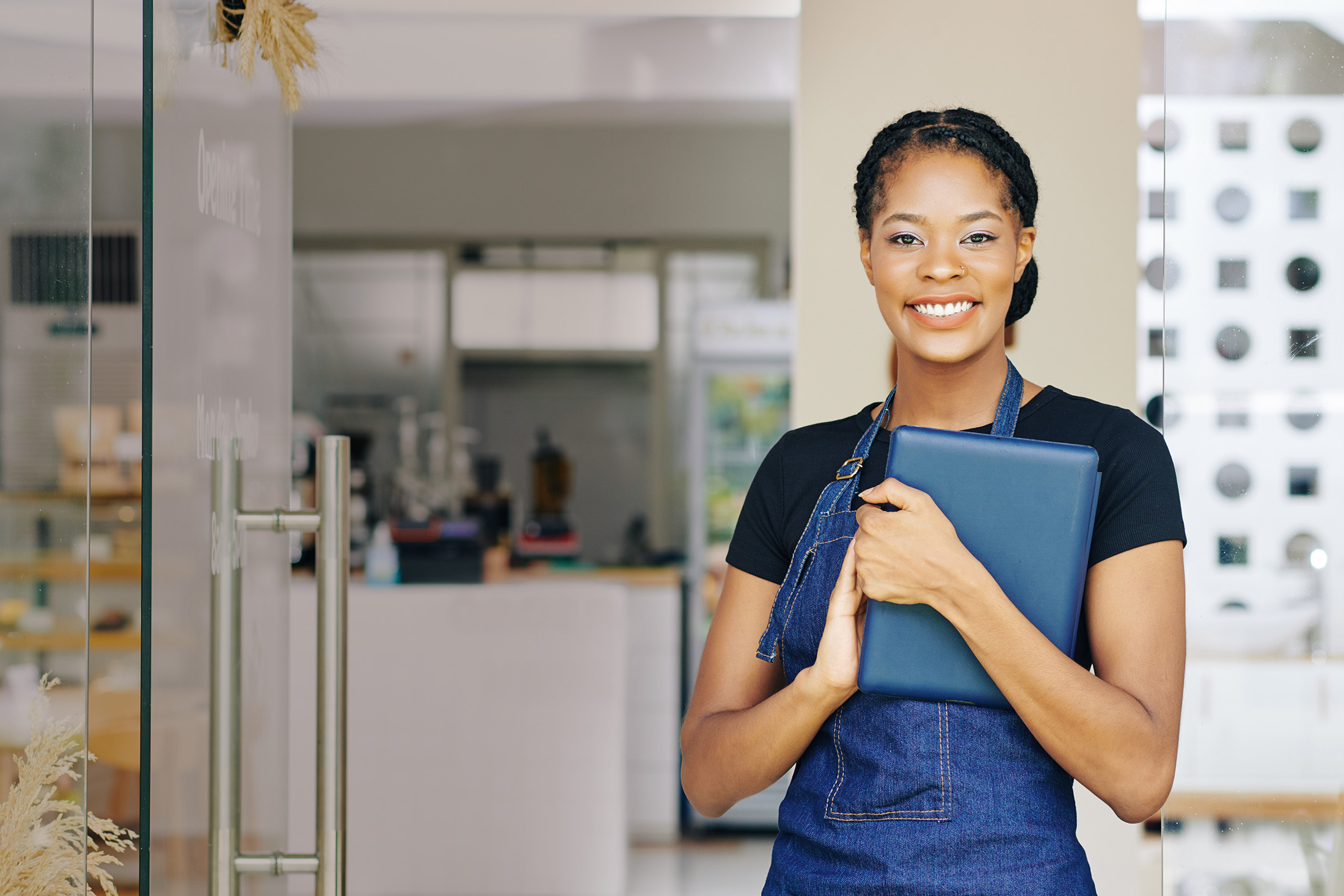 Portrait of pretty smiling young Black woman with tablet computer in hands standing in doors of coffeeshop she just opened